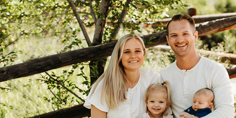 Family of four sitting outdoors in front of a wooden fence and greenery; two adults in white shirts, one holding a baby in a blue blanket, with a child between them.