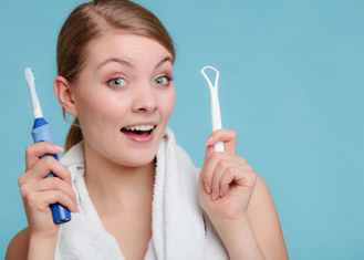 A person holds an electric toothbrush in the left hand and a white tongue scraper in the right, with a white towel draped around the neck. The solid light blue background and visible tools emphasize oral hygiene, creating a clean, informative visual suitable for dental education or product promotion.