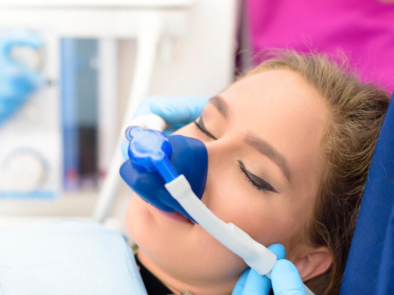A patient lies back in a dental chair while a gloved hand approaches their mouth, indicating an ongoing clinical procedure. The surrounding instruments, blue glove, and containers establish a sterile and professional dental setting.