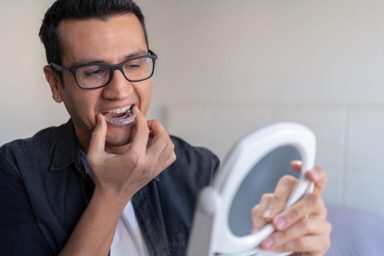 A man wearing glasses carefully inserts a clear dental mouthguard using a small mirror for guidance, demonstrating a precise orthodontic routine.