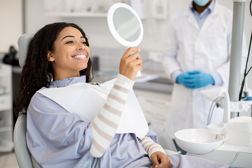 A patient sits in a dental chair wearing a bib, holding a round mirror to check their teeth, while a dentist in a white coat and blue gloves stands nearby in a clinic setting with dental equipment visible.