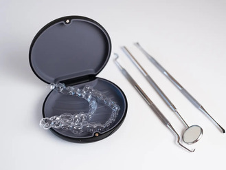 Clear dental aligner in black case beside mirror, probe, and scaler on white surface.